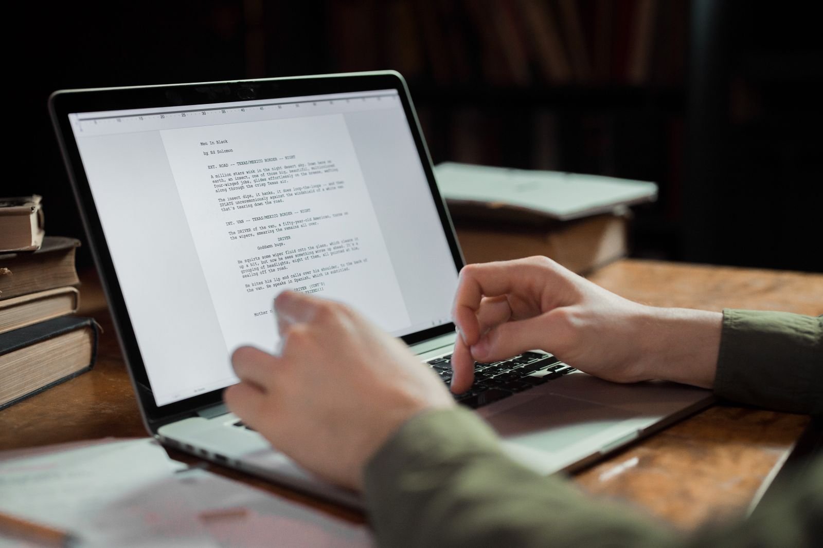 Hands typing on a laptop showing a long text document, with marked-up pages beside it